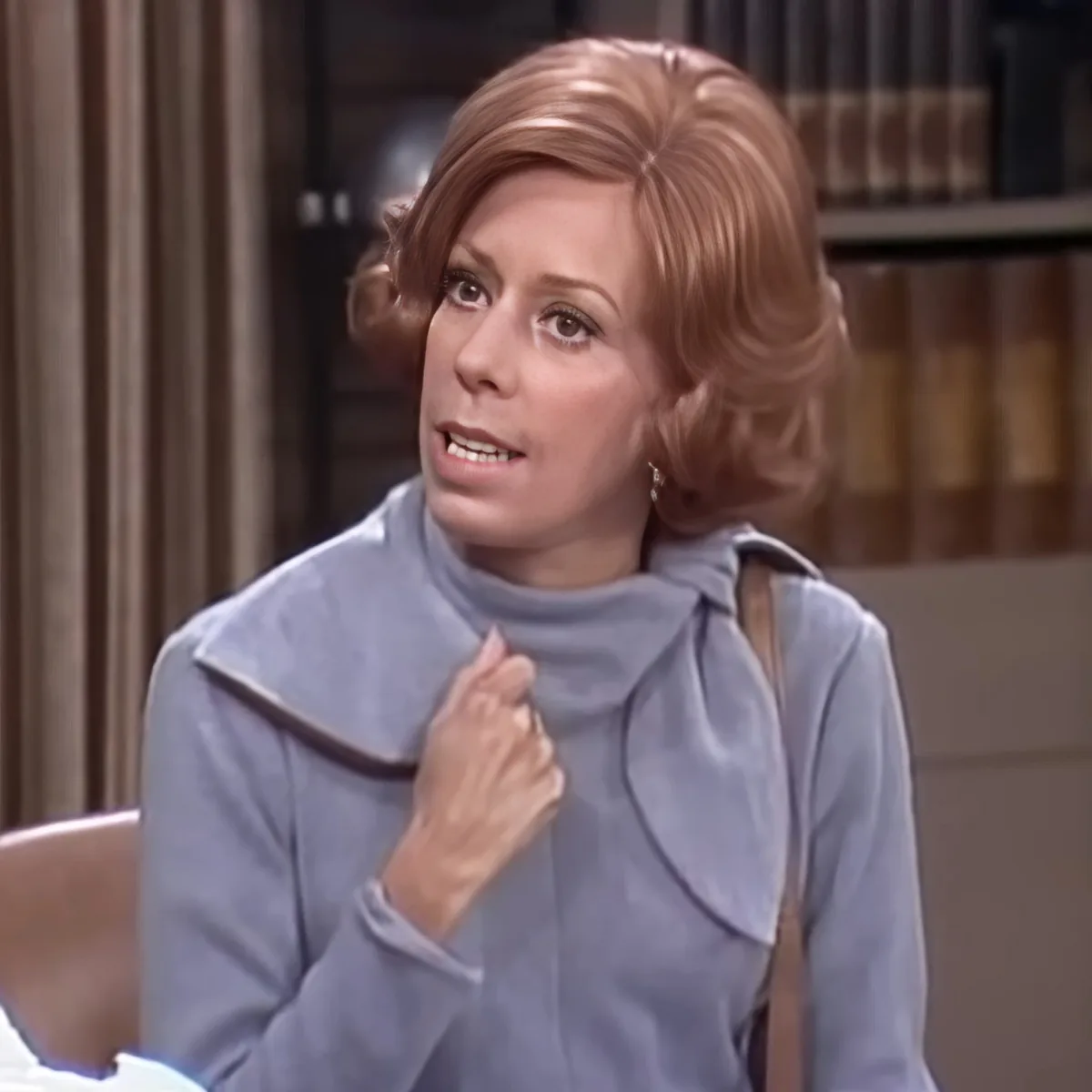 A woman with short, styled hair, wearing a light blue turtleneck and a brown strap, is adjusting her clothing in a room...