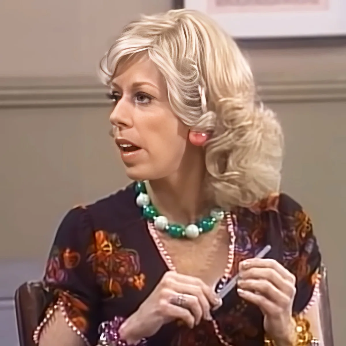 A woman with blonde hair, wearing a floral dress and a green and white beaded necklace, is holding a small object in her...