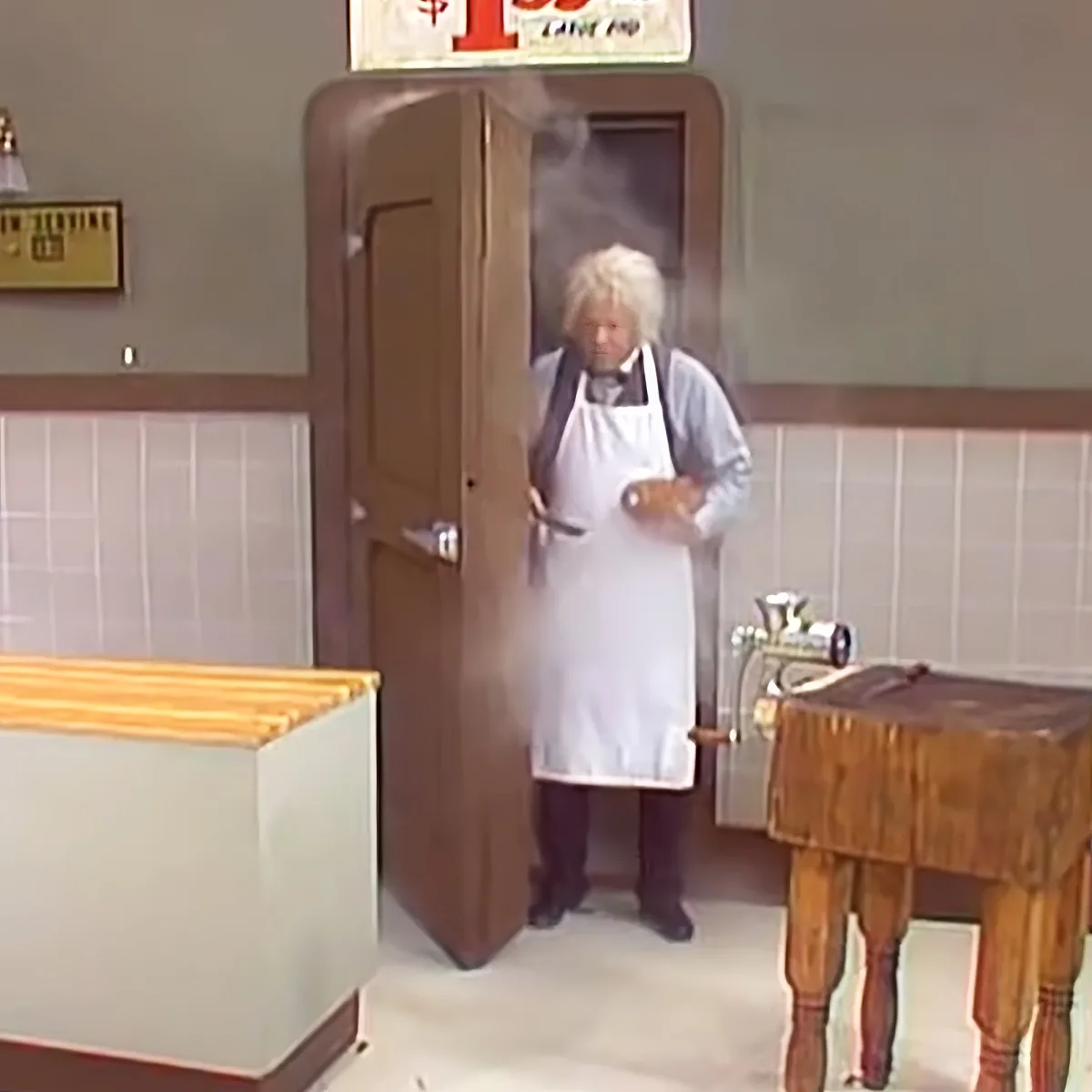 A chef in a white apron stands in a kitchen, steam rising from a pot, holding a knife and a container.