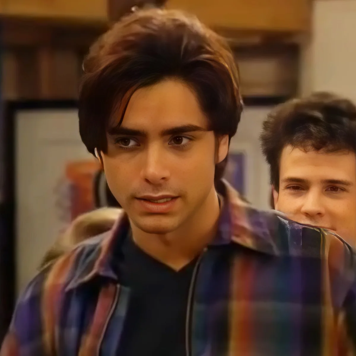 A man with short brown hair and a plaid shirt looks to the side, with another person partially visible behind him.