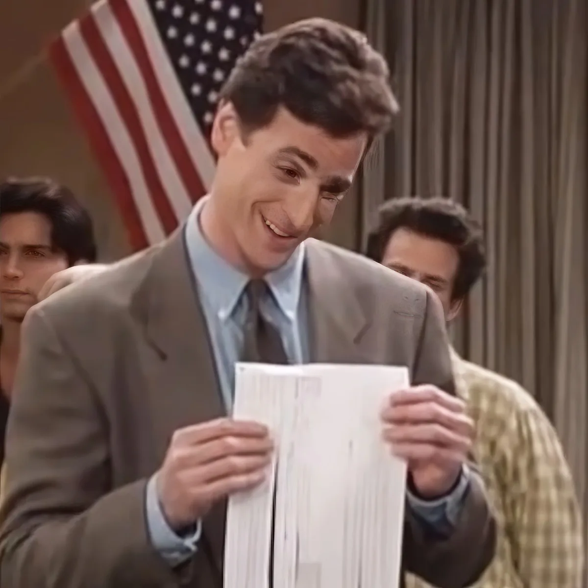 A man in a suit smiles while holding a stack of papers, with an American flag and other people in the background.