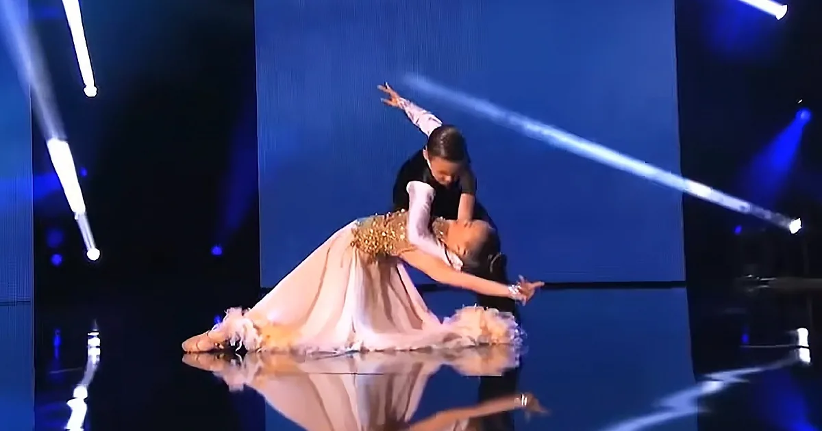 A ballerina in a pink dress and gold embellishments is performing a dramatic pose on stage with a partner.