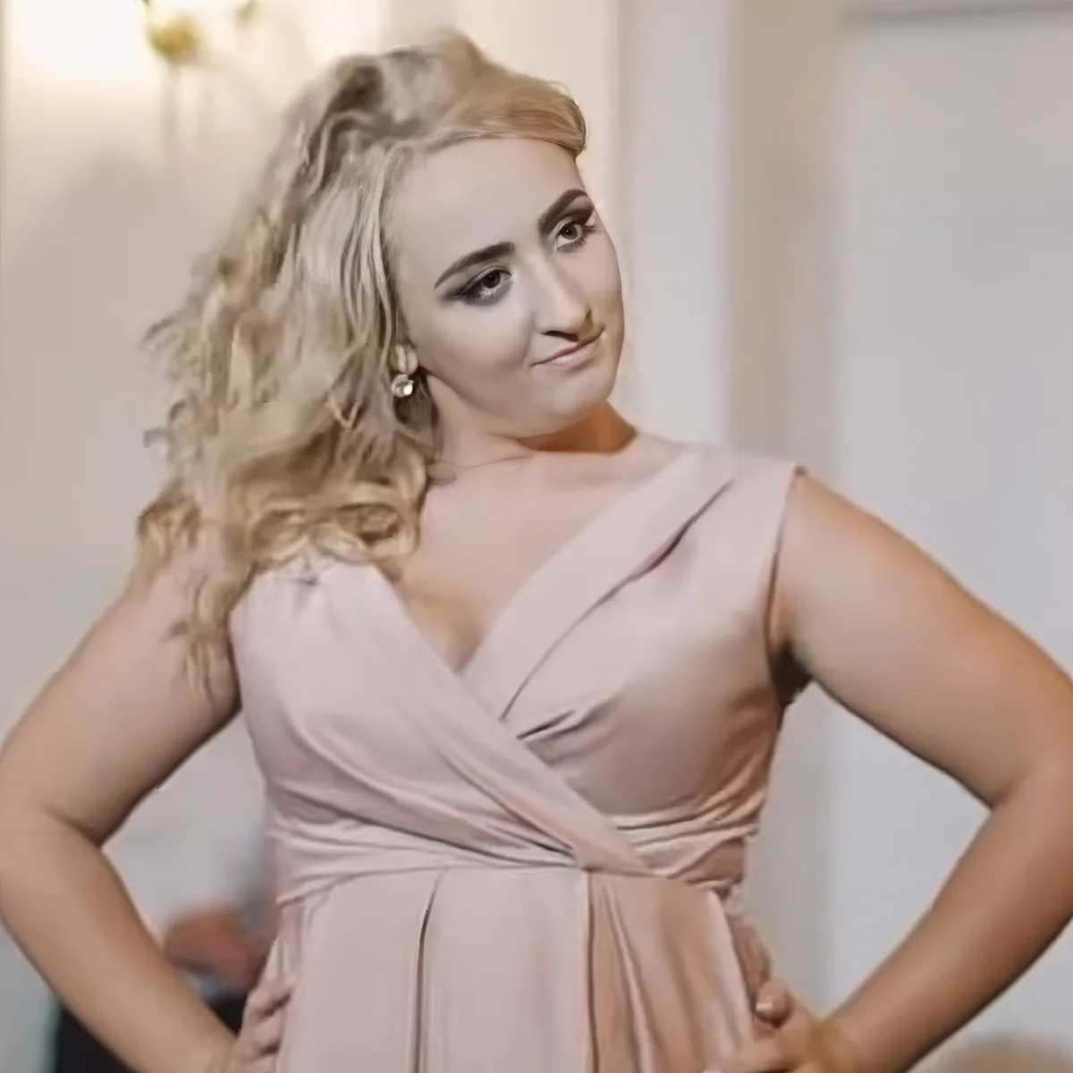 A woman with blonde curls in a pink dress stands with hands on hips, smiling.