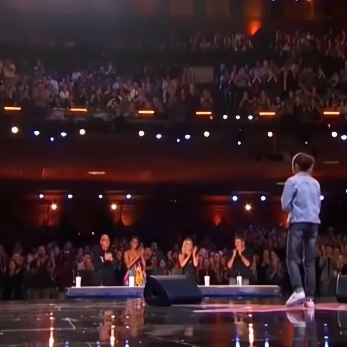 A man stands on stage, facing an audience applauding in a dimly lit venue.