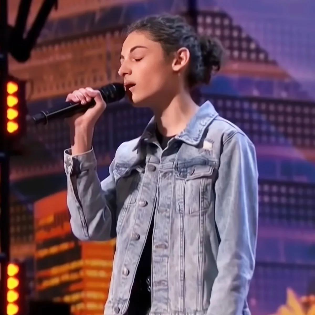 A person in a denim jacket sings into a microphone on a stage with a cityscape backdrop.