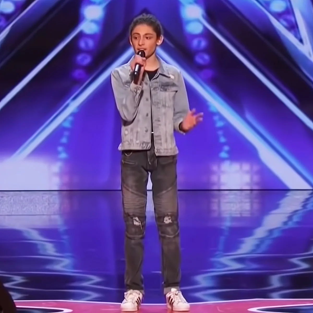 A young person in a denim jacket and ripped jeans stands on stage holding a microphone, gesturing with one hand.