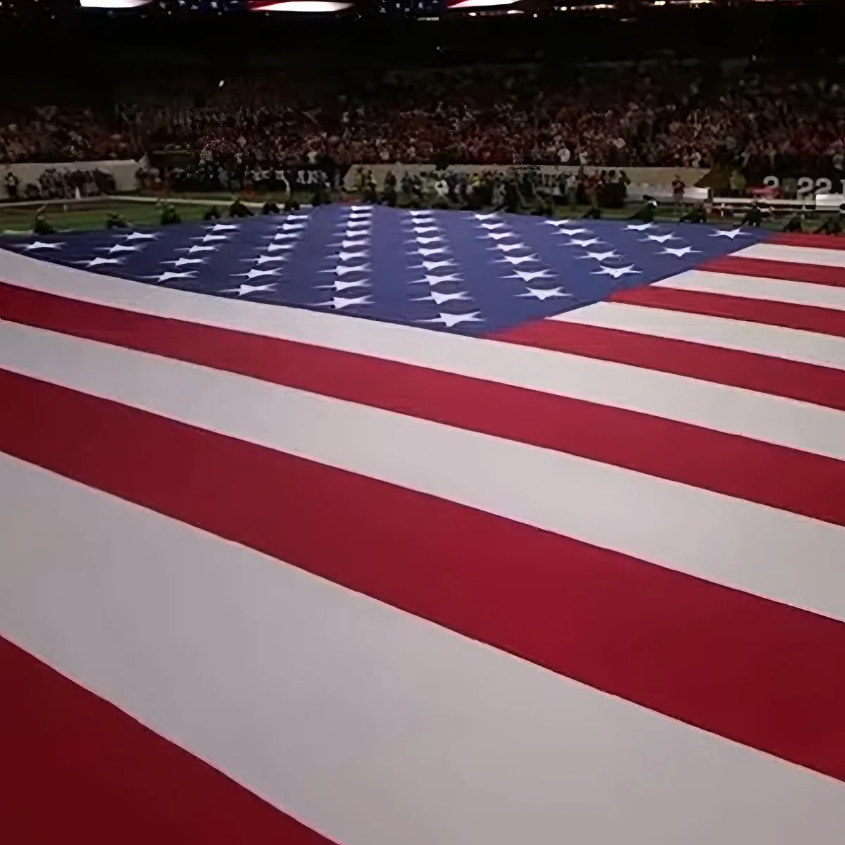 A large American flag is displayed prominently in a stadium with spectators in the background.
