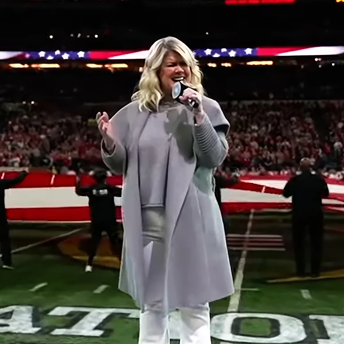 A woman in a gray coat sings into a microphone on a football field with an American flag in the background.
