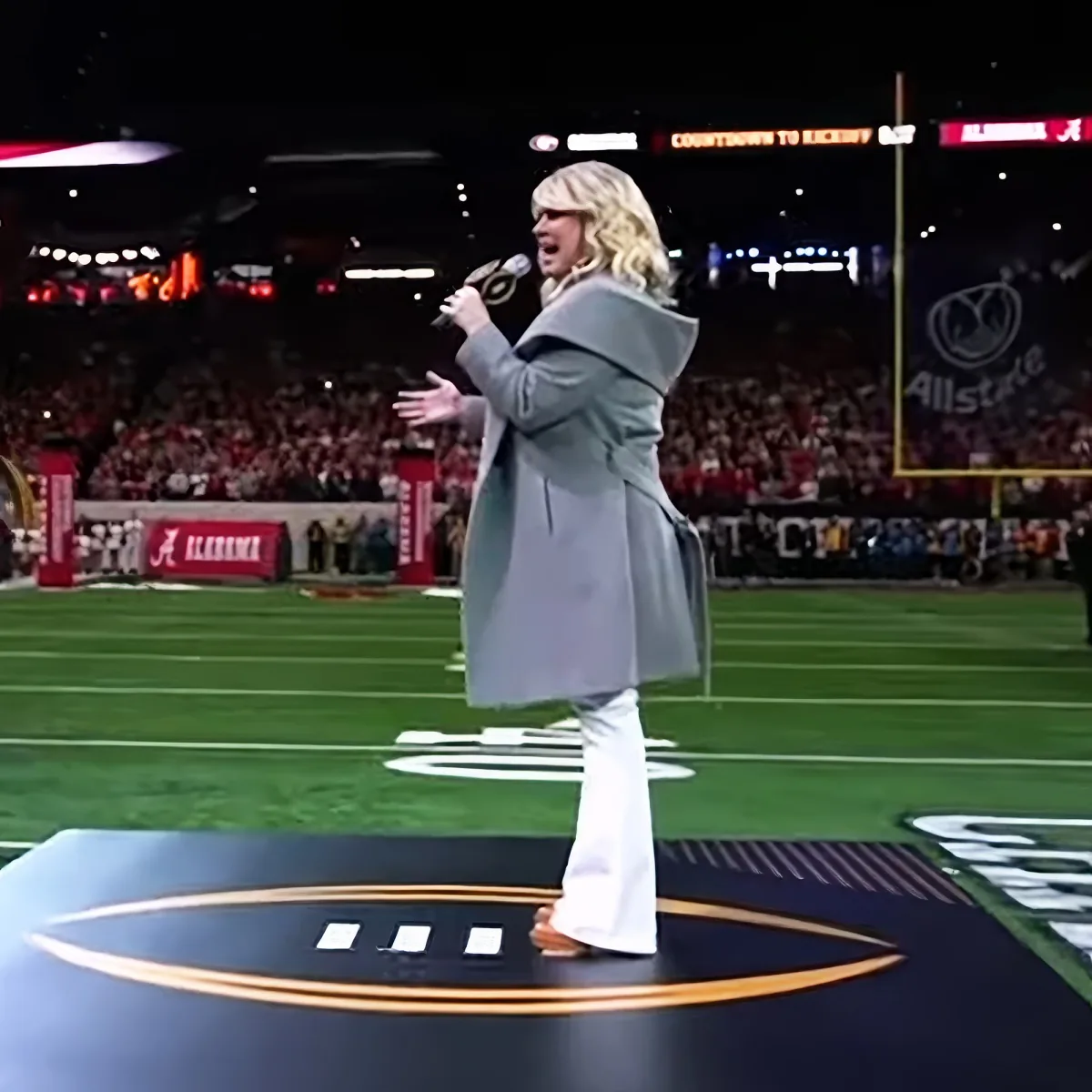 A woman in a gray coat sings on a football field with a crowd and stadium lights in the background.