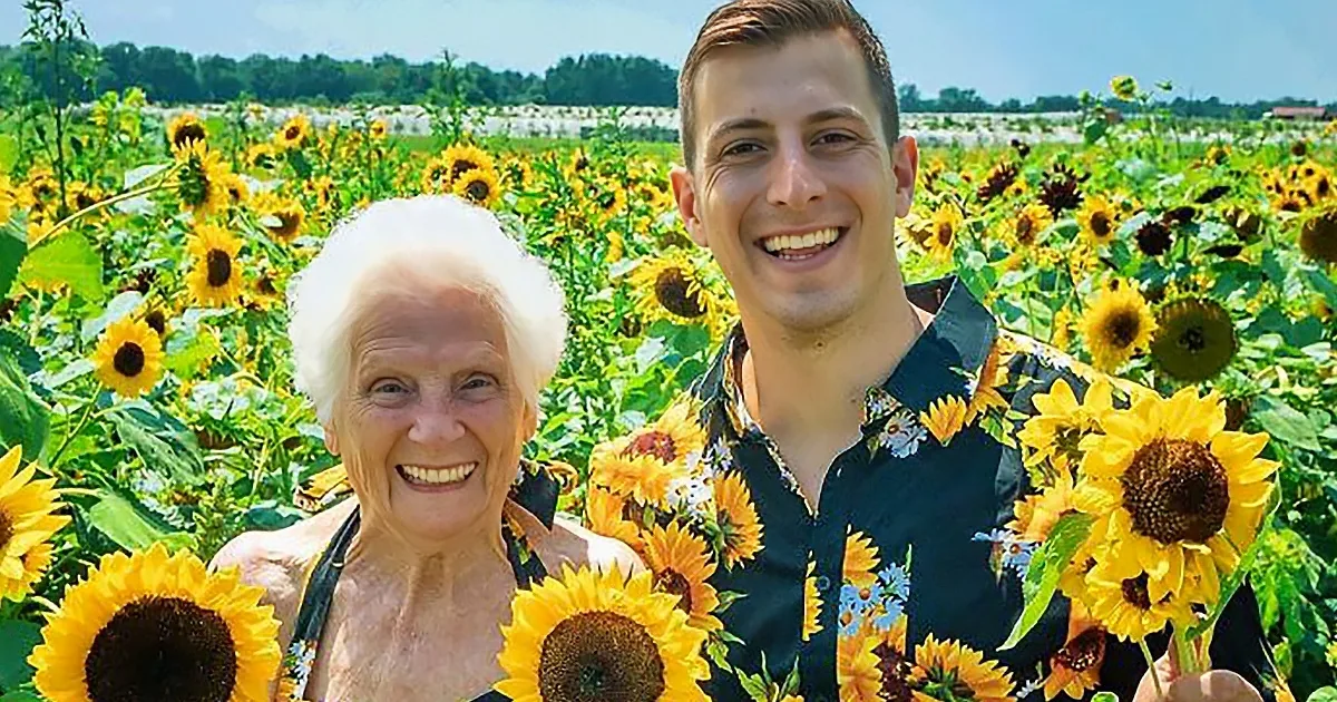 A smiling elderly woman and a young man stand amidst a vibrant sunflower field, enjoying a sunny day.