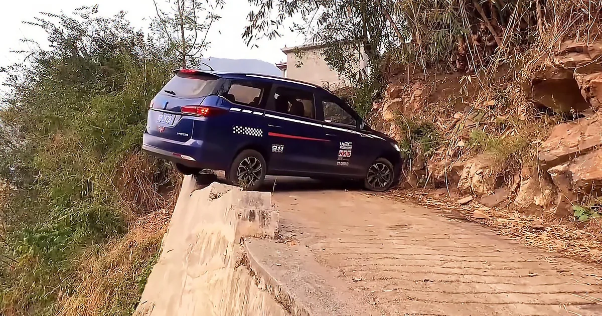A blue car is driving on a narrow dirt path surrounded by rocks and vegetation.
