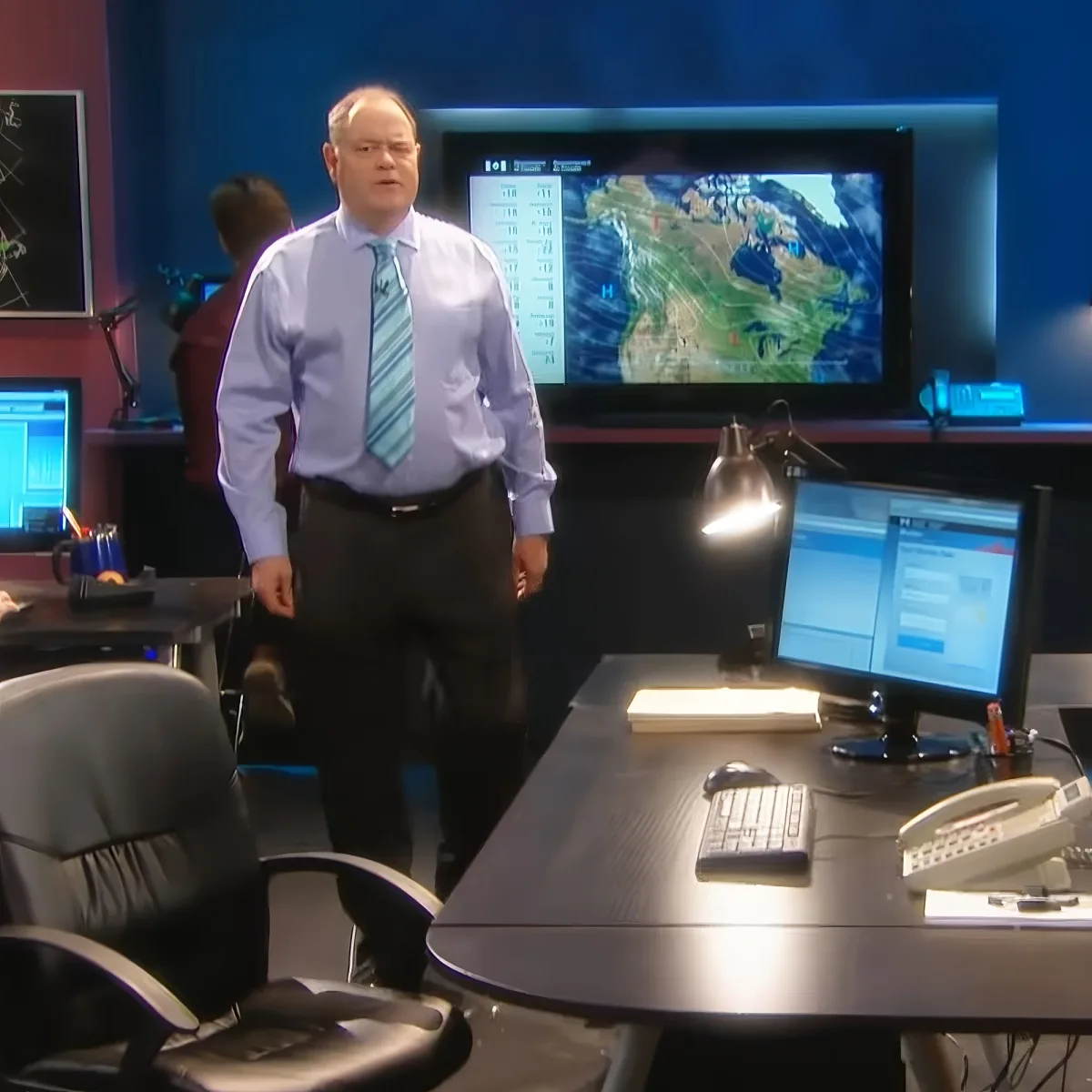 A man in a light blue shirt and tie stands in a weather forecast studio with a map on the screen behind him.