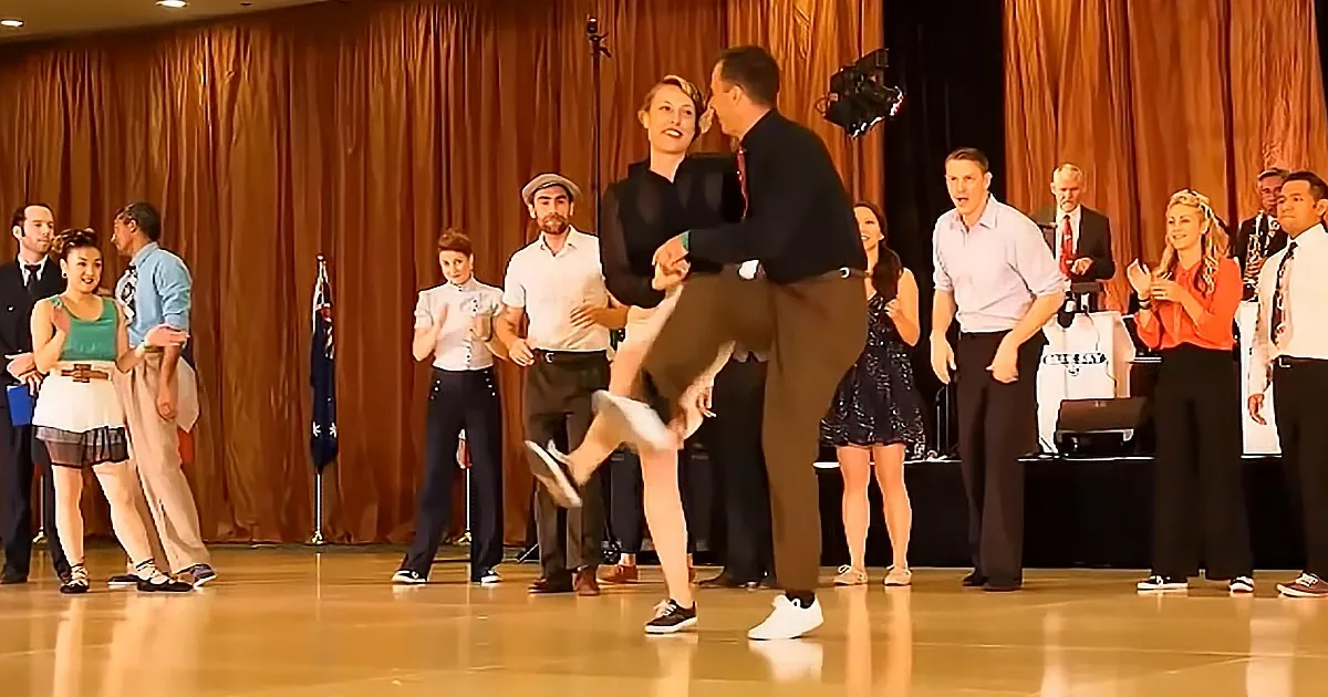 A man and woman are dancing on stage, with the man lifting the woman's leg in a dynamic pose.