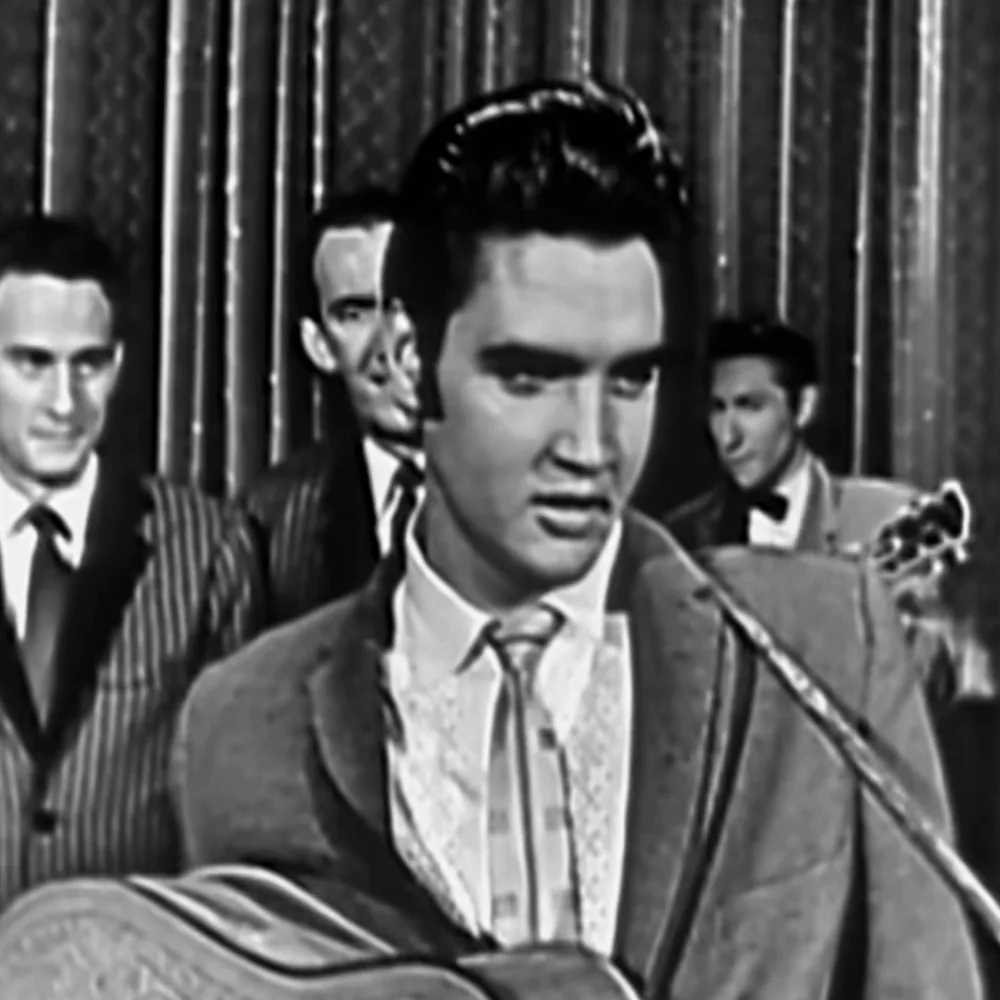 Elvis Presley, in a suit, sings into a microphone with a band behind him.