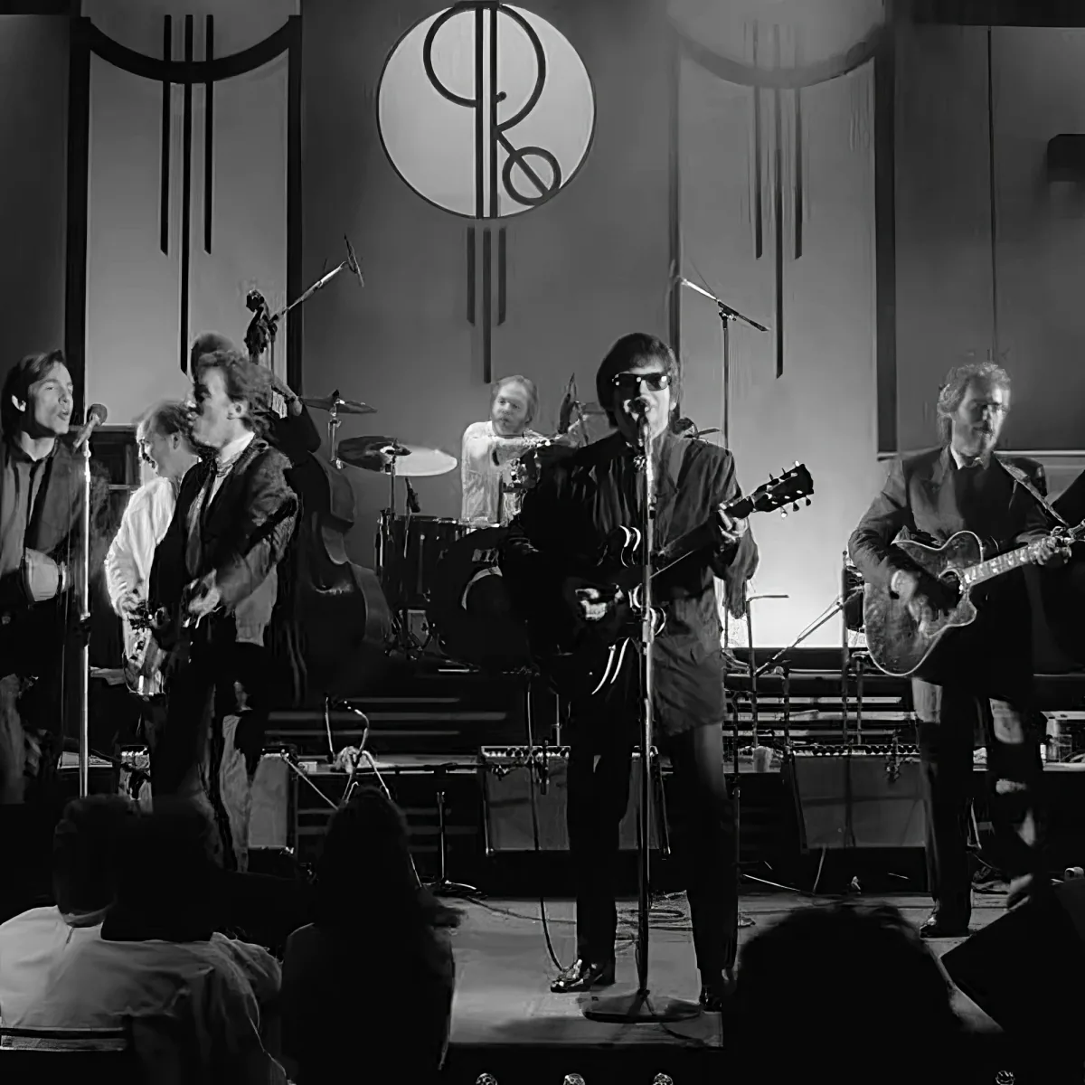 A man in sunglasses plays guitar on stage with a band, surrounded by microphones and instruments.