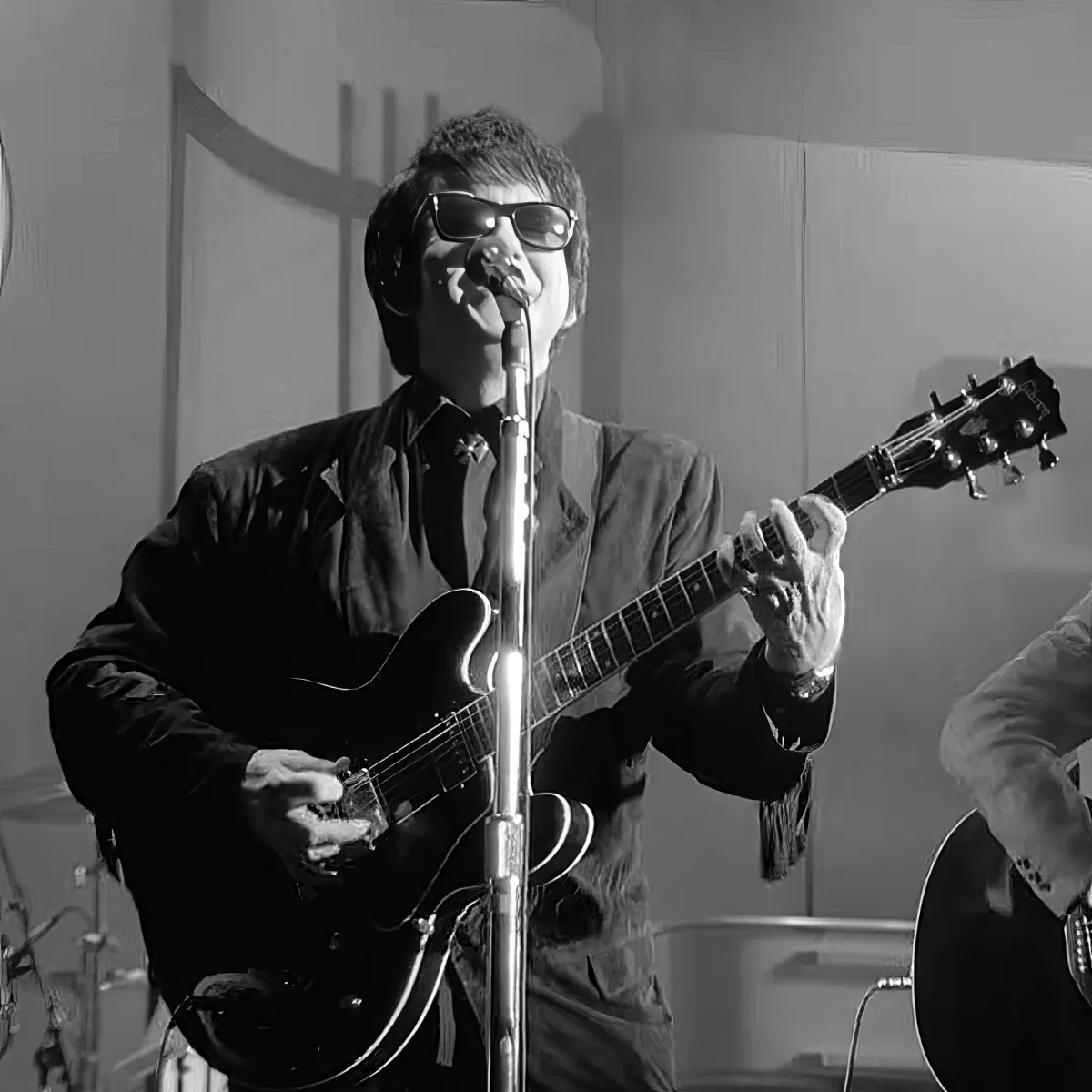 A person in a suit plays a guitar and sings into a microphone, wearing sunglasses.