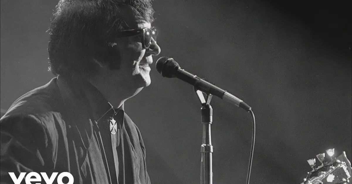A person wearing glasses sings into a microphone on stage, illuminated by stage lighting.