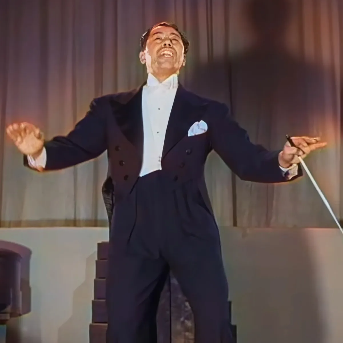 A man in a tuxedo and bow tie is joyfully singing on stage, holding a cane.