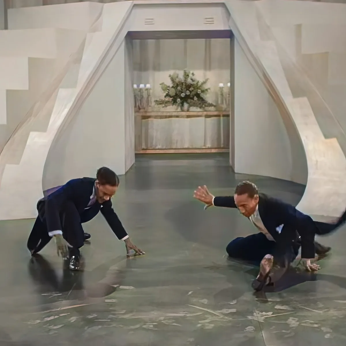 Two men in suits crouch on a marble floor, one reaching out as if to help the other.