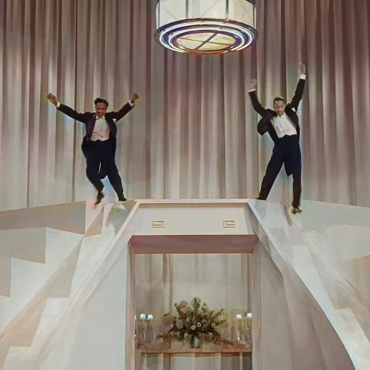 Two men in suits jump joyfully on a stage with a chandelier and floral arrangement below.