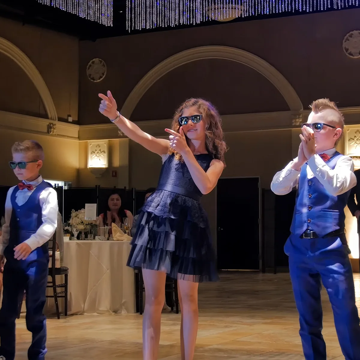 A girl in a blue dress and sunglasses points while two boys in matching outfits clap in a formal setting.
