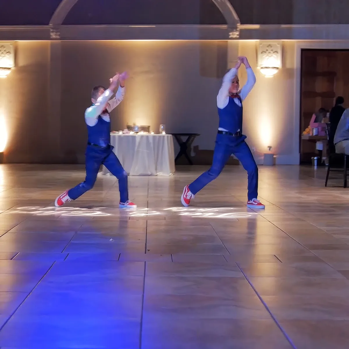 Two individuals in blue outfits and red sneakers dance on a wooden floor with a table and chairs in the background.