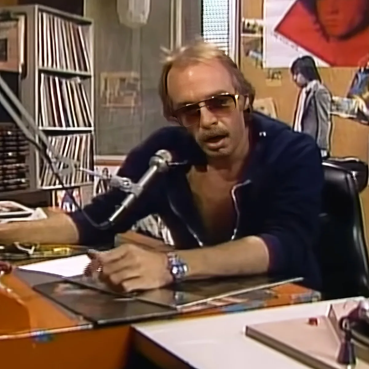 A man in sunglasses sits at a desk with a microphone, surrounded by records and posters.