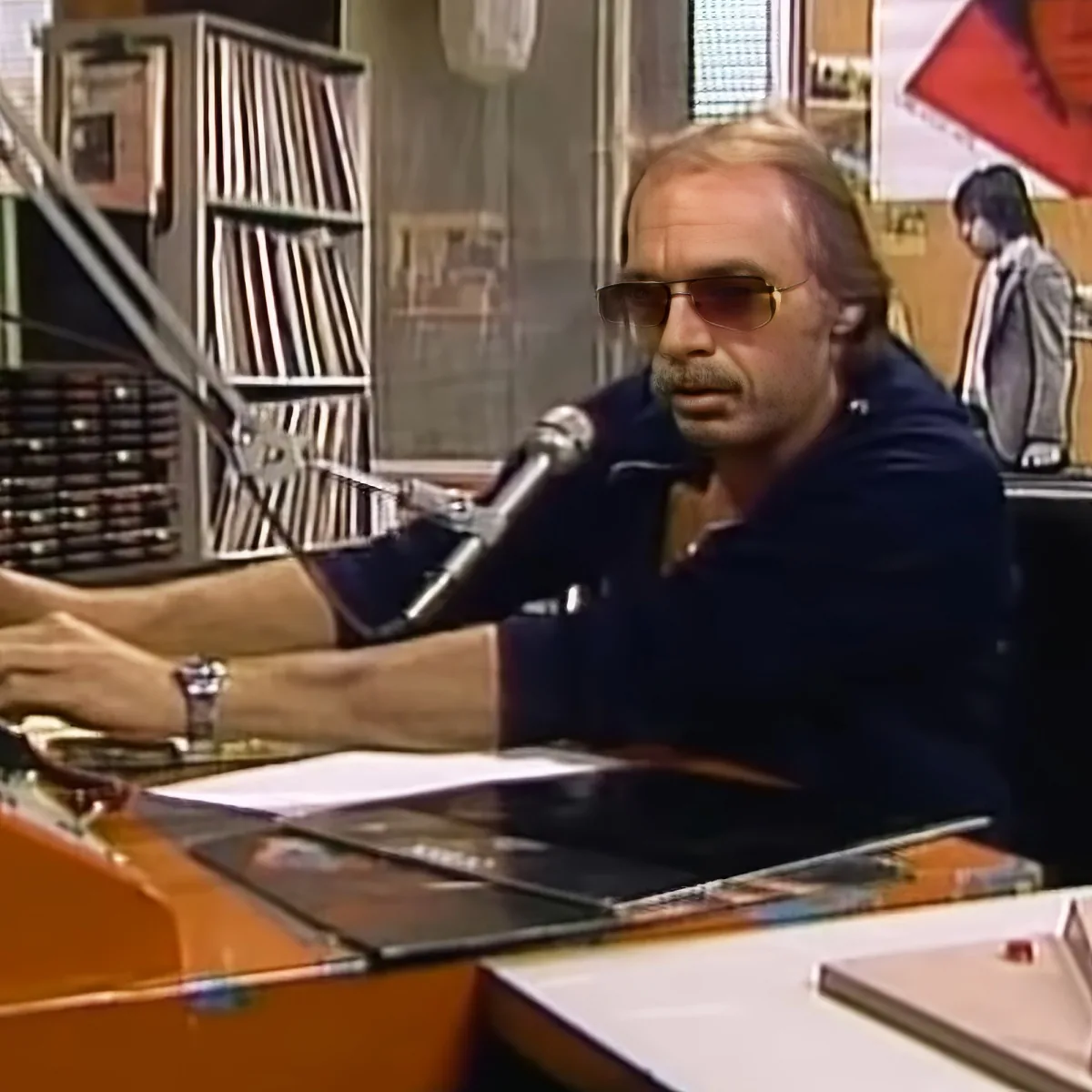 A man in sunglasses sits at a desk with a microphone, surrounded by papers and a record player, in a radio studio.