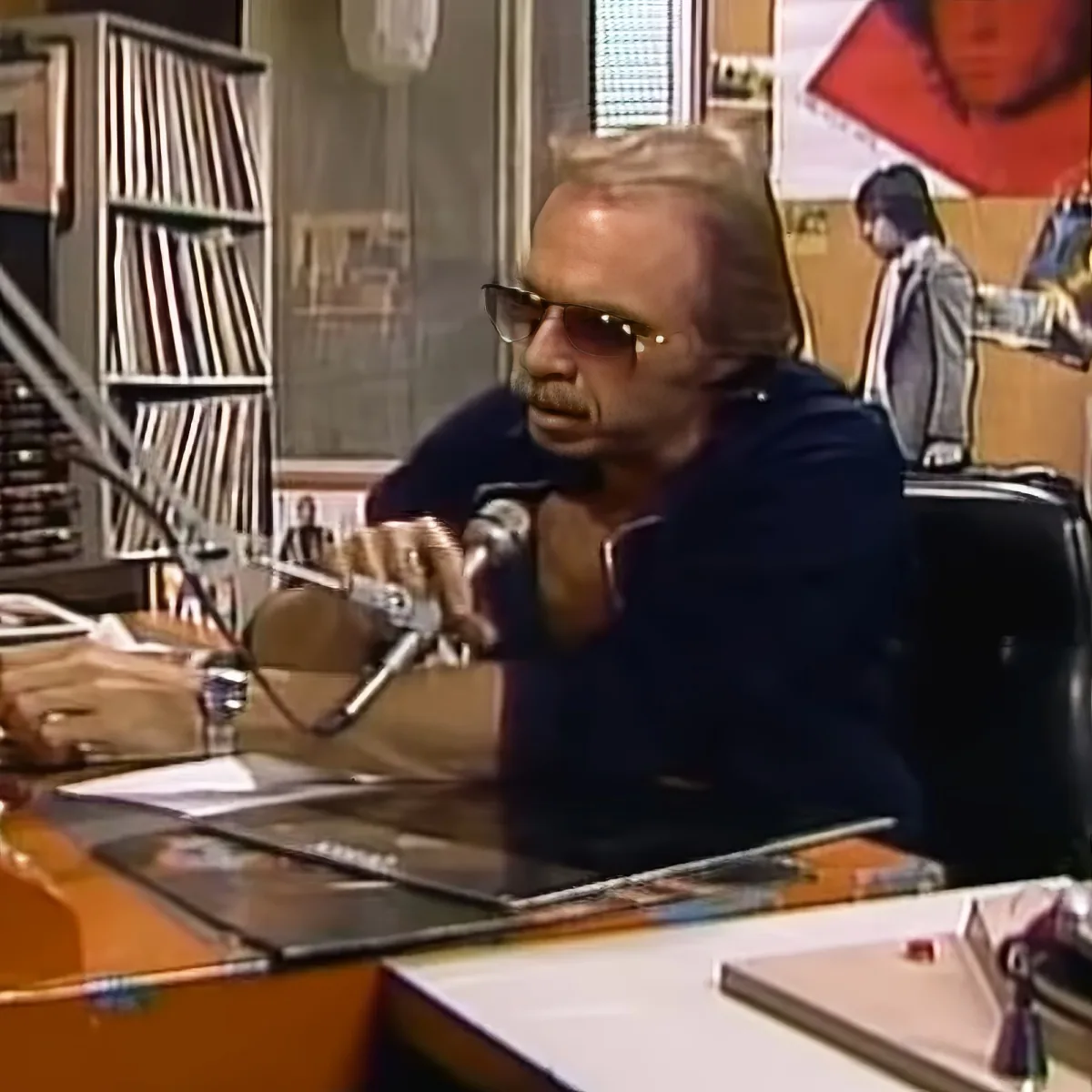 A man wearing sunglasses sits at a desk with a microphone, surrounded by books and posters.