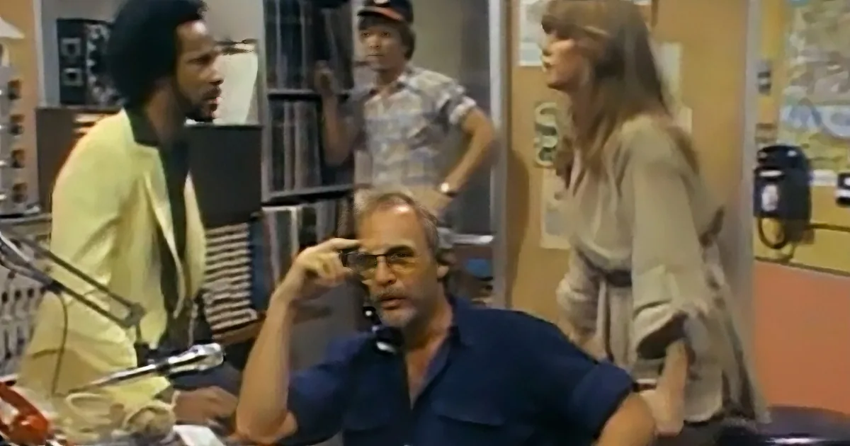 A man in a blue shirt sits at a desk, holding a phone to his ear, with others in the background.