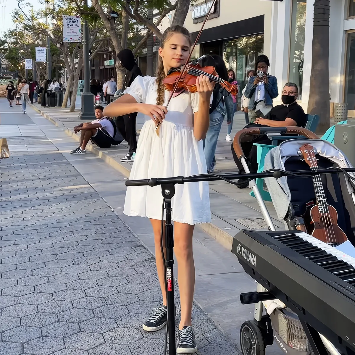 A girl in a white dress plays violin on a sidewalk with a Yamaha keyboard and ukulele nearby.