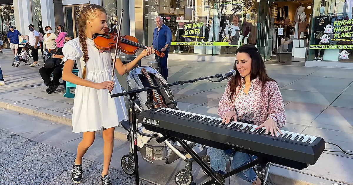 A young girl plays violin while a woman plays keyboard in a public setting, with a stroller and passersby in the background.