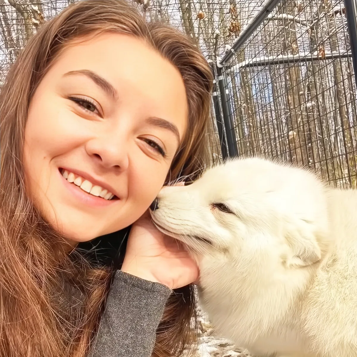 A smiling woman with long brown hair holds a white dog, which is licking her hand.
