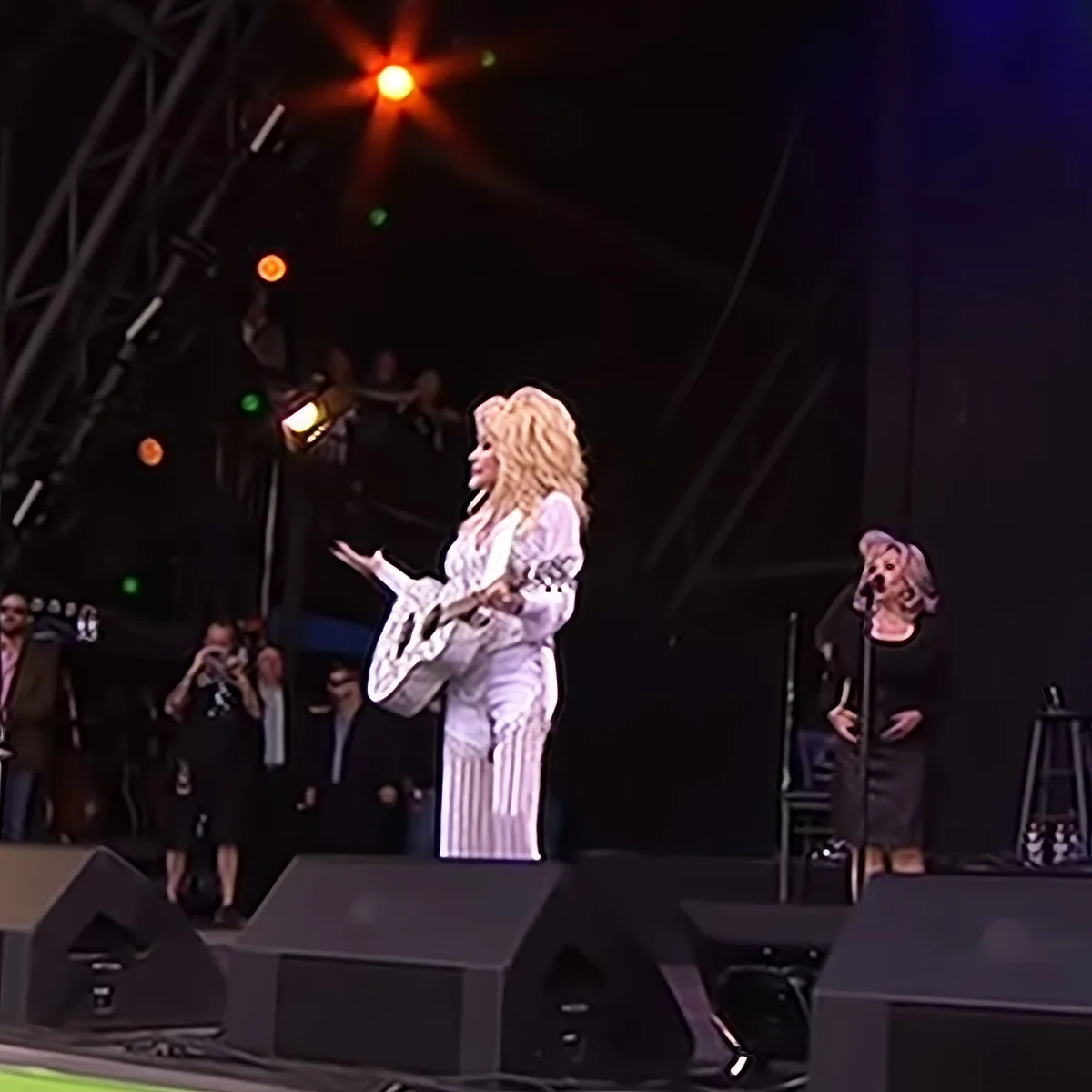 A woman in a white dress plays guitar on stage, with another woman singing nearby, surrounded by bright stage lights.