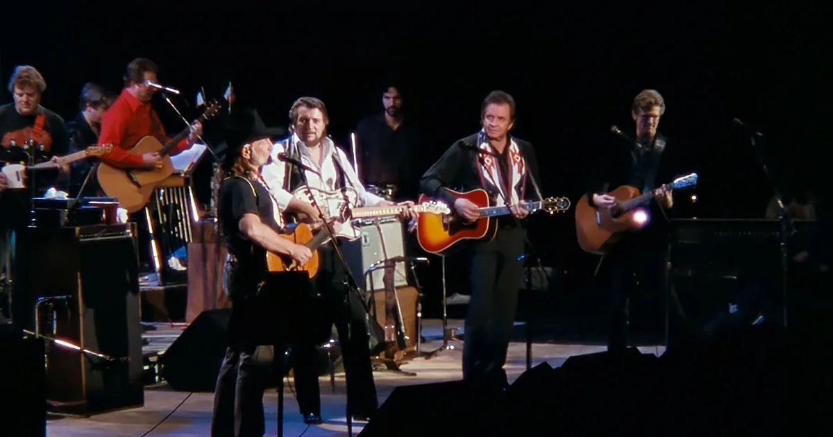 A man in a cowboy hat plays guitar on stage with a band, surrounded by other musicians.