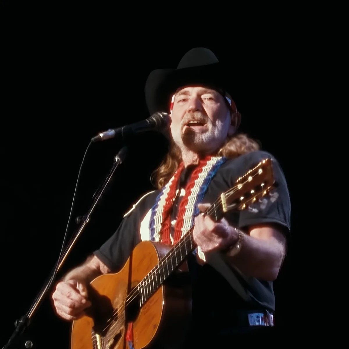 A man in a cowboy hat plays an acoustic guitar on stage, singing into a microphone.