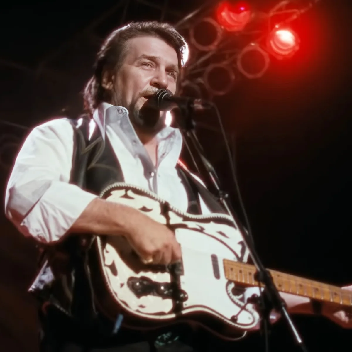 A man in a white shirt and black vest plays a white electric guitar on stage with a microphone.
