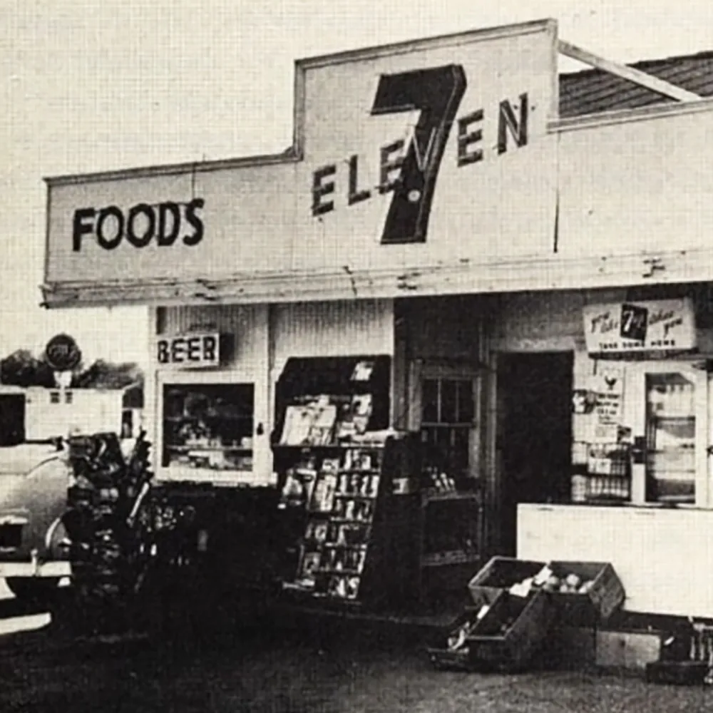 An old 7-Eleven store with "Foods" and "Beer" signs, vintage cars parked, and merchandise displayed outside.