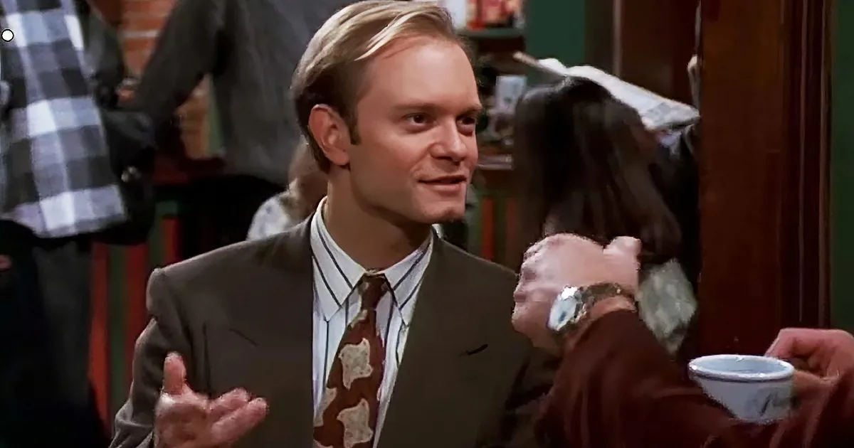 A man in a suit and tie gestures while holding a cup, surrounded by people in a social setting.