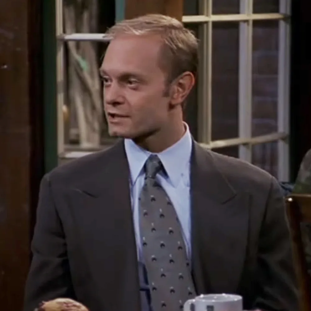 A man in a suit and tie is speaking, with a window and door in the background.