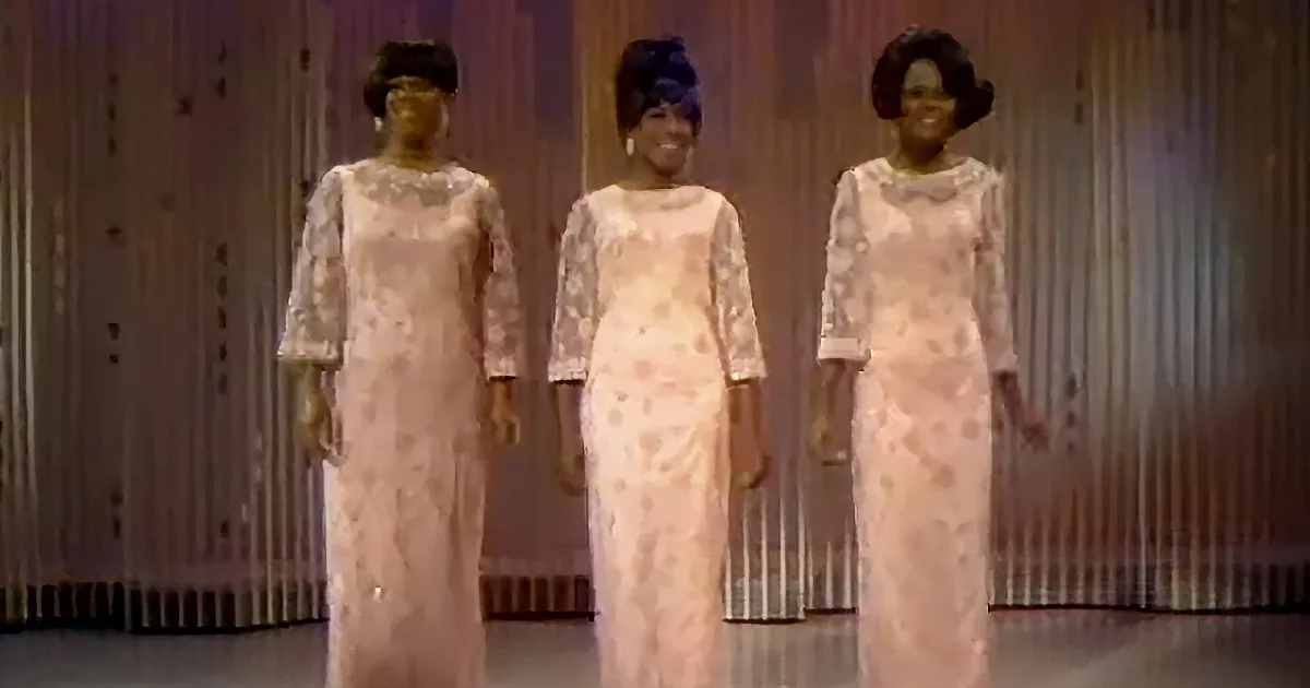 Three women in matching white lace dresses stand side by side on a stage.