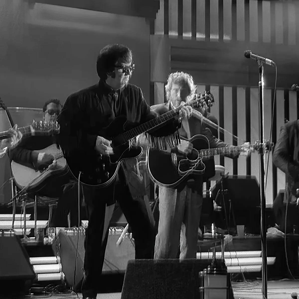 A man in glasses plays an electric guitar on stage, surrounded by other musicians.