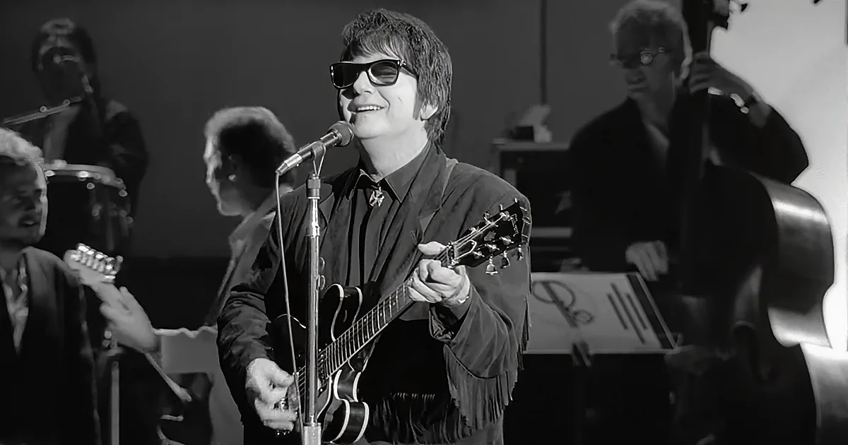A musician in sunglasses plays guitar on stage, smiling, with a band in the background.