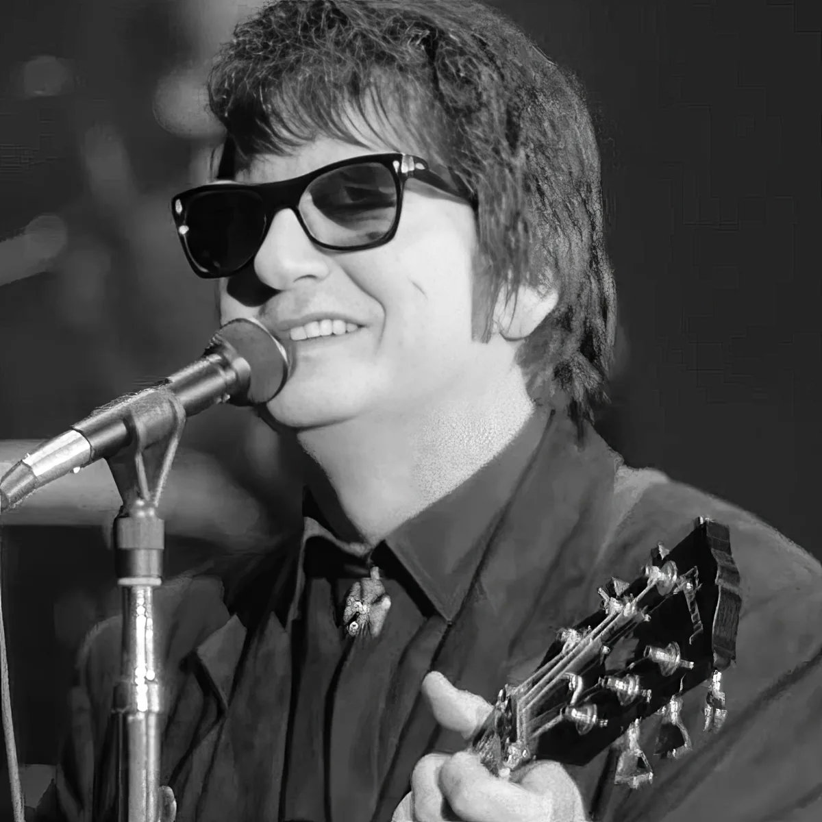 A person wearing sunglasses and a black shirt sings into a microphone while holding a guitar.