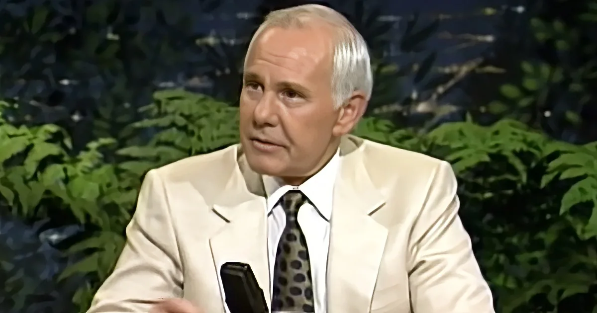 A man in a white suit and patterned tie is speaking, with a green leafy background.