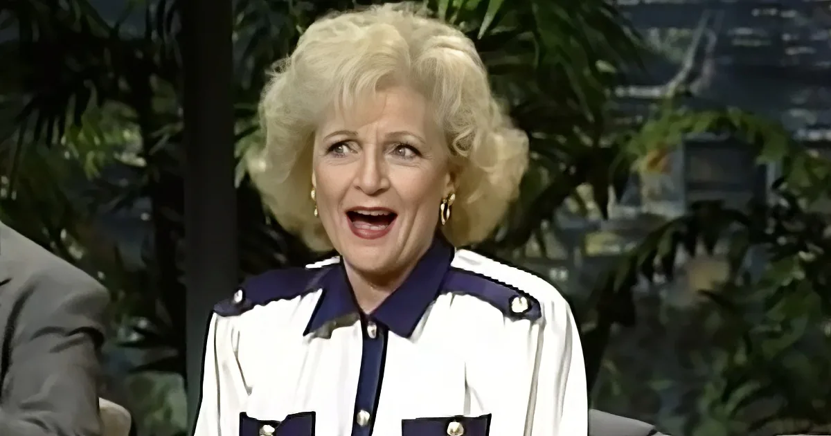 A woman with blonde hair, wearing a white and blue outfit, is smiling and appears to be on a talk show set with plants in...