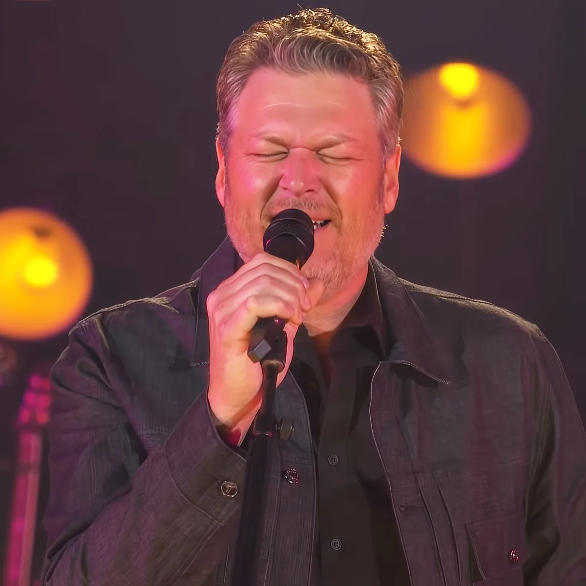 A man with a beard sings into a microphone, wearing a dark shirt, with a warm light in the background.