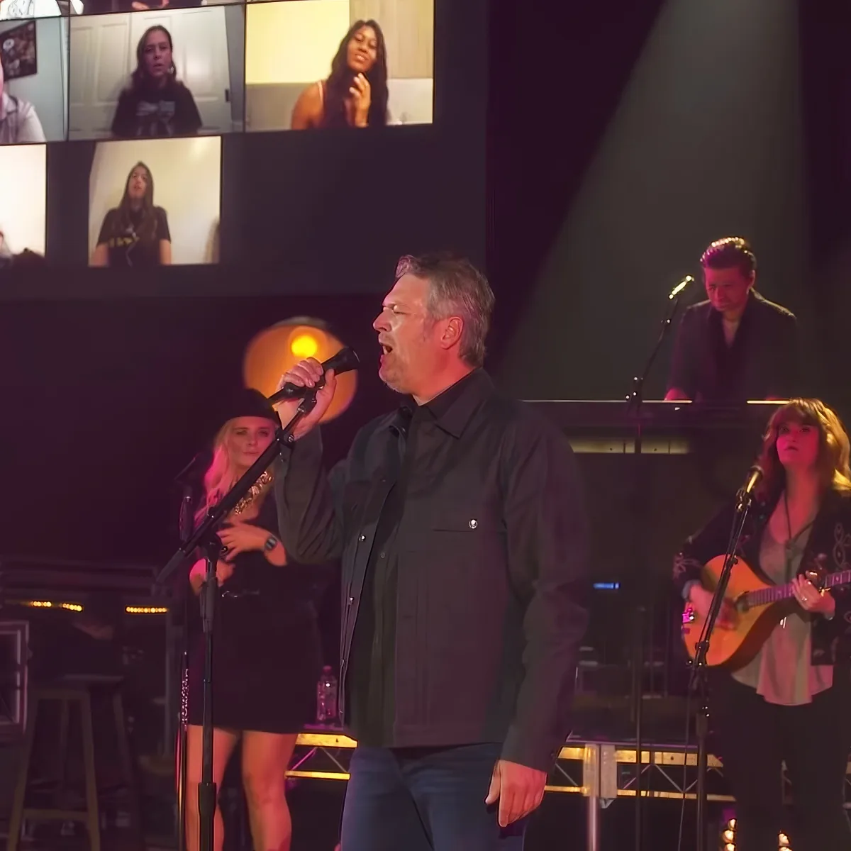 A man sings into a microphone on stage with a band and a virtual audience displayed on screens behind him.