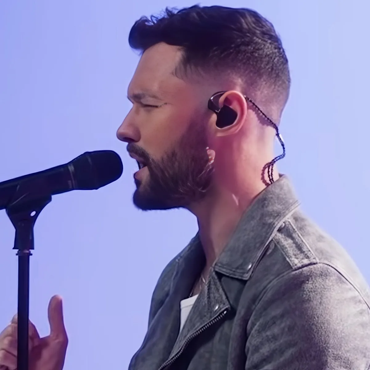A man with a beard and earpiece sings into a microphone, wearing a denim jacket.
