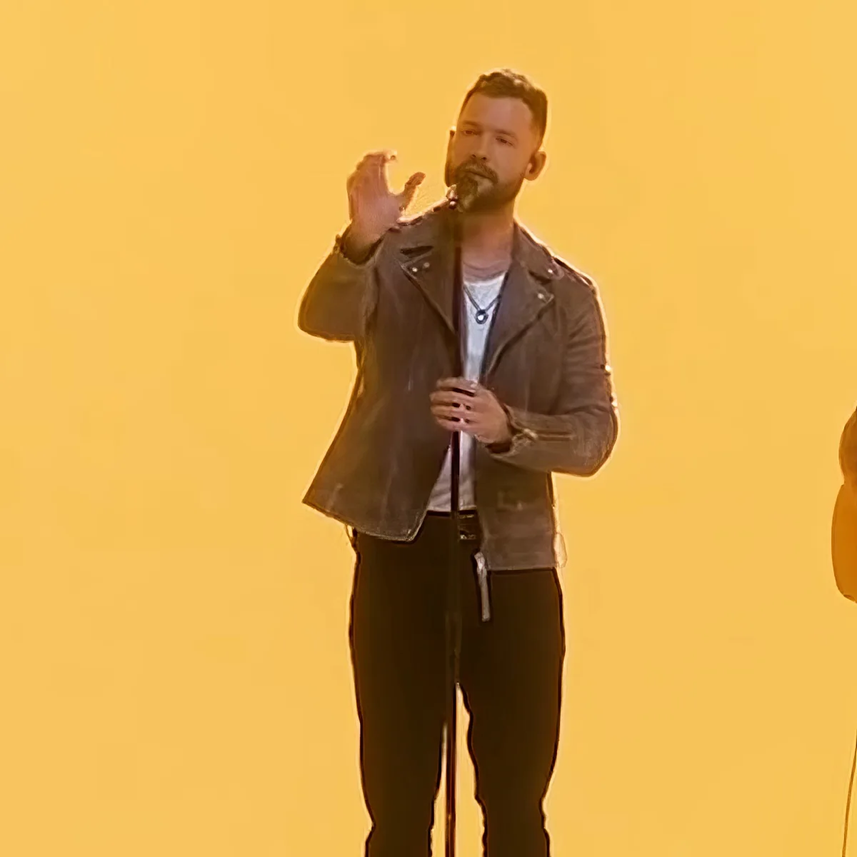 A man in a leather jacket and jeans stands on stage, holding a microphone, with a yellow background.